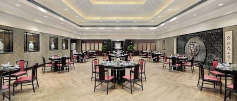 A spacious dining area with multiple round tables and chairs, a high ceiling with recessed lighting, and traditional decor elements.