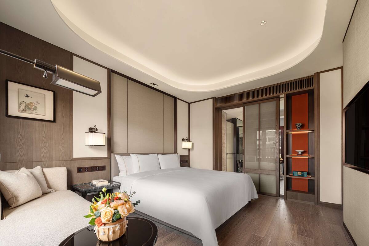 A modern hotel room with a large bed, a desk, a chair, a nightstand, a wall-mounted lamp, a framed artwork, and a decorative vase with flowers.