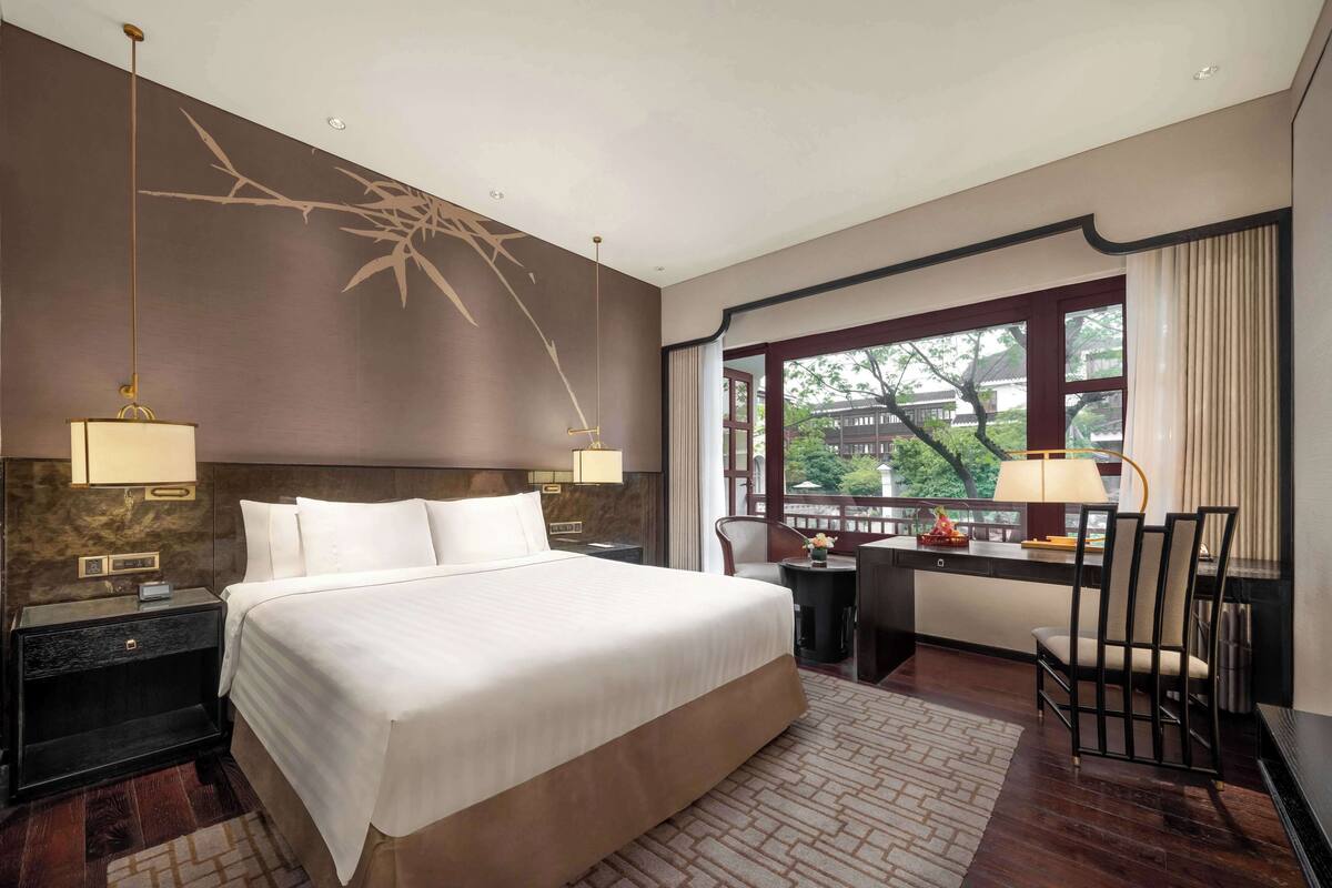 A hotel room with a large bed, a desk with a chair, a window with a view of greenery, and a decorative wall hanging.