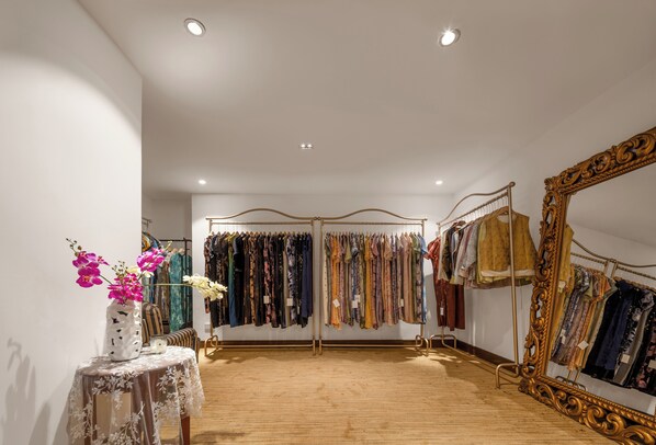 A clothing store with racks of clothes, a large ornate mirror, and a table with a vase of flowers.