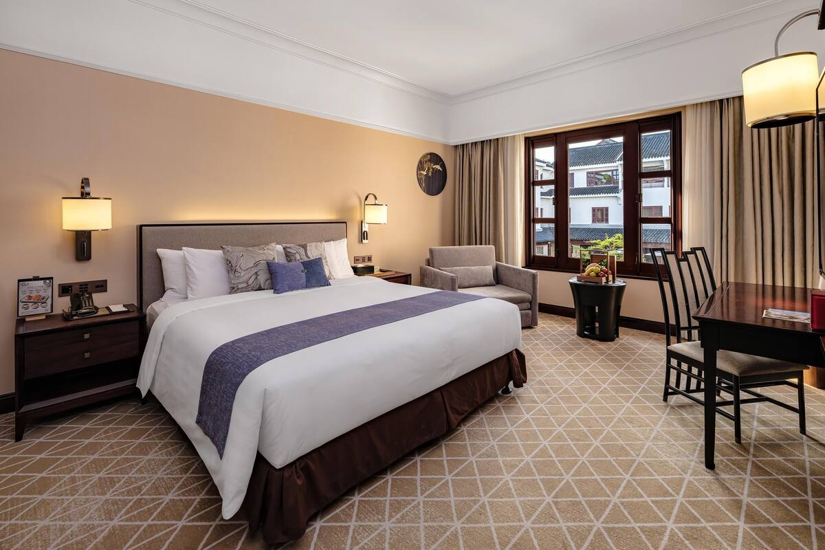 A hotel room with a large bed, a desk, and a chair. There are two bedside tables, each with a lamp. A window with curtains is visible, and a clock is on the wall.