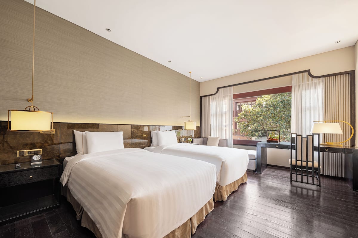 A hotel room with two beds, a desk, and a view of greenery outside.