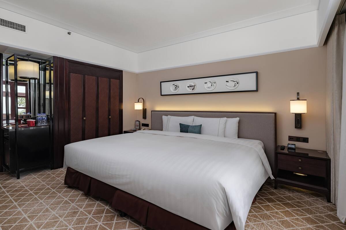 A hotel room with a large bed, a desk with a chair, a wardrobe, and a wall-mounted artwork.