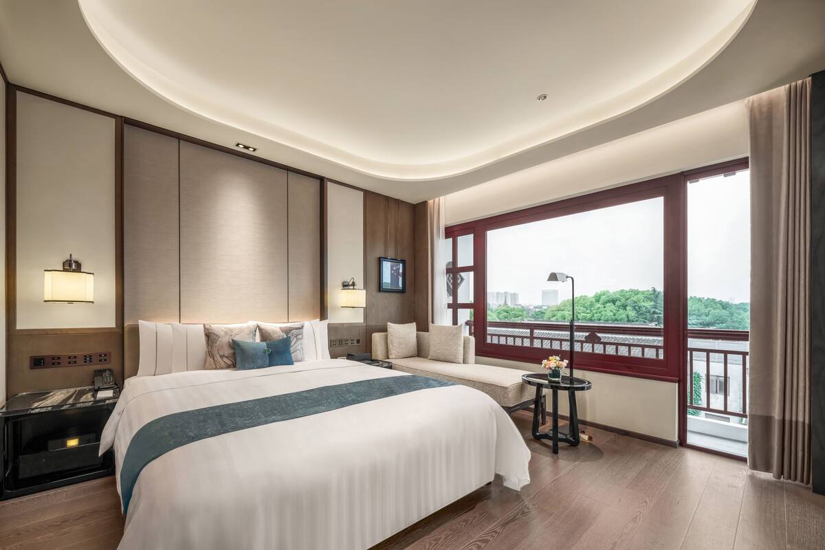 A hotel room with a large bed, a sofa, a TV, a desk, and a balcony with a view.