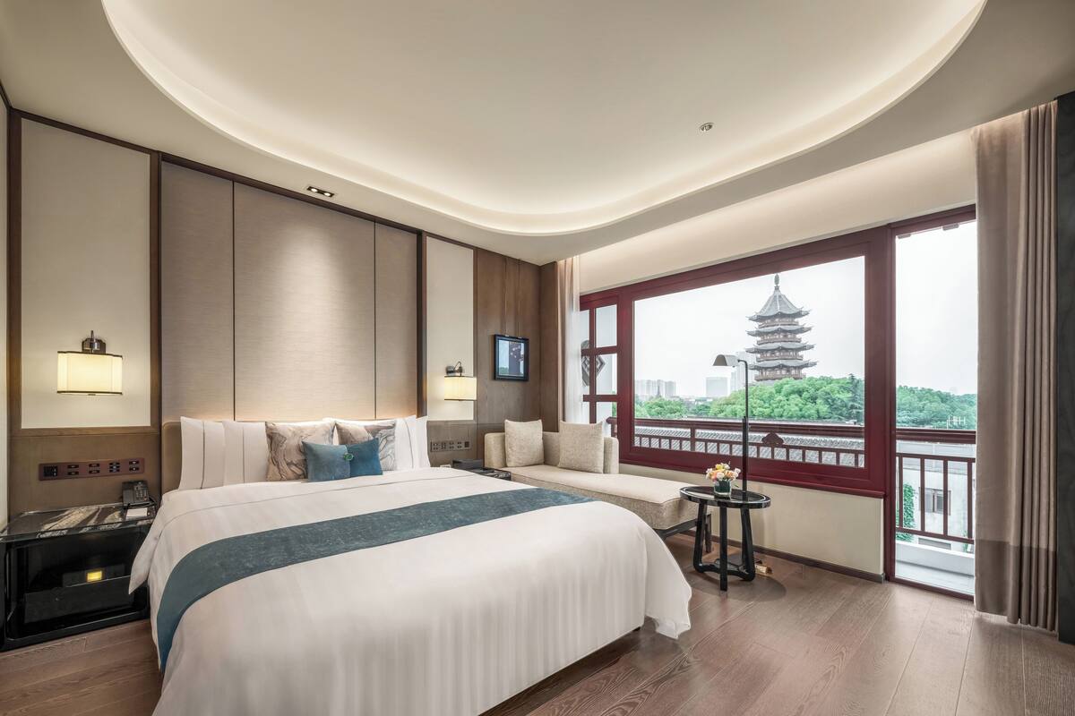 A hotel room with a large bed, a bedside table, a TV, a window with a view of a pagoda, and a balcony.