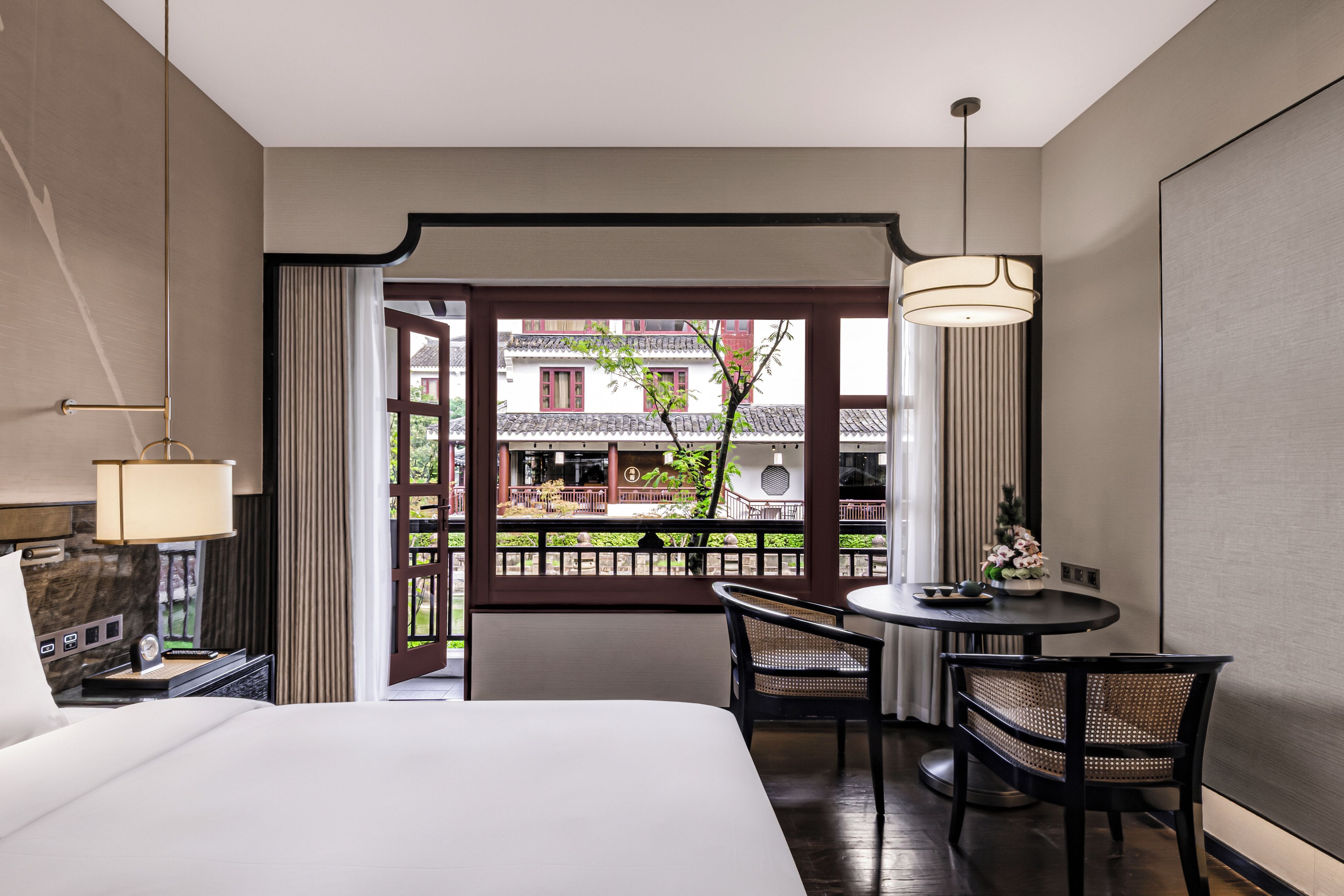 A hotel room with a bed, a table with chairs, a lamp, and a view of a traditional building through an open door.