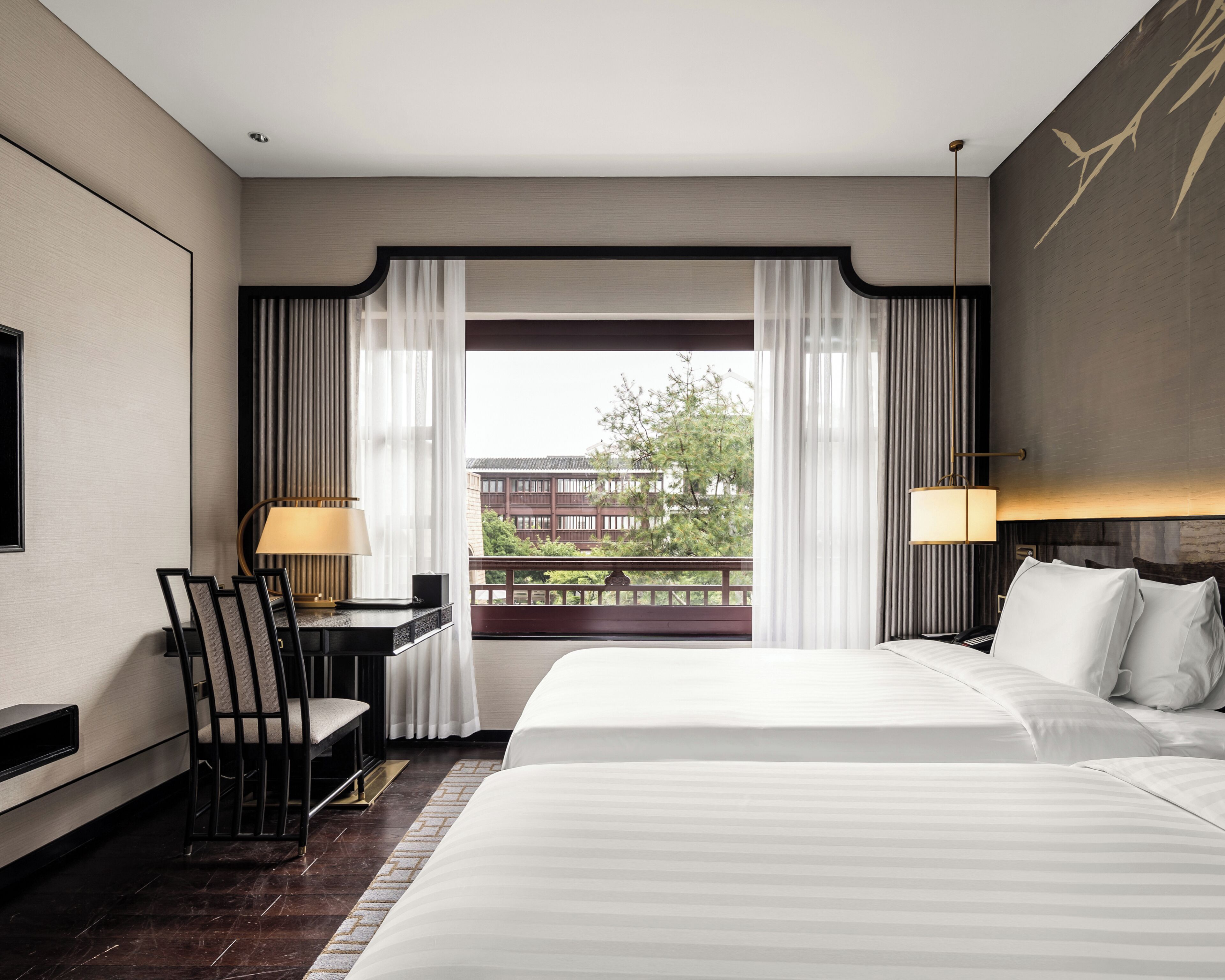 A hotel room with a large bed, a desk with a chair, a lamp, and a view of a building and trees through sheer curtains.
