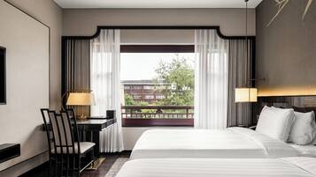 A hotel room with a large bed, a desk with a chair, a lamp, and a view of a building and trees through sheer curtains.