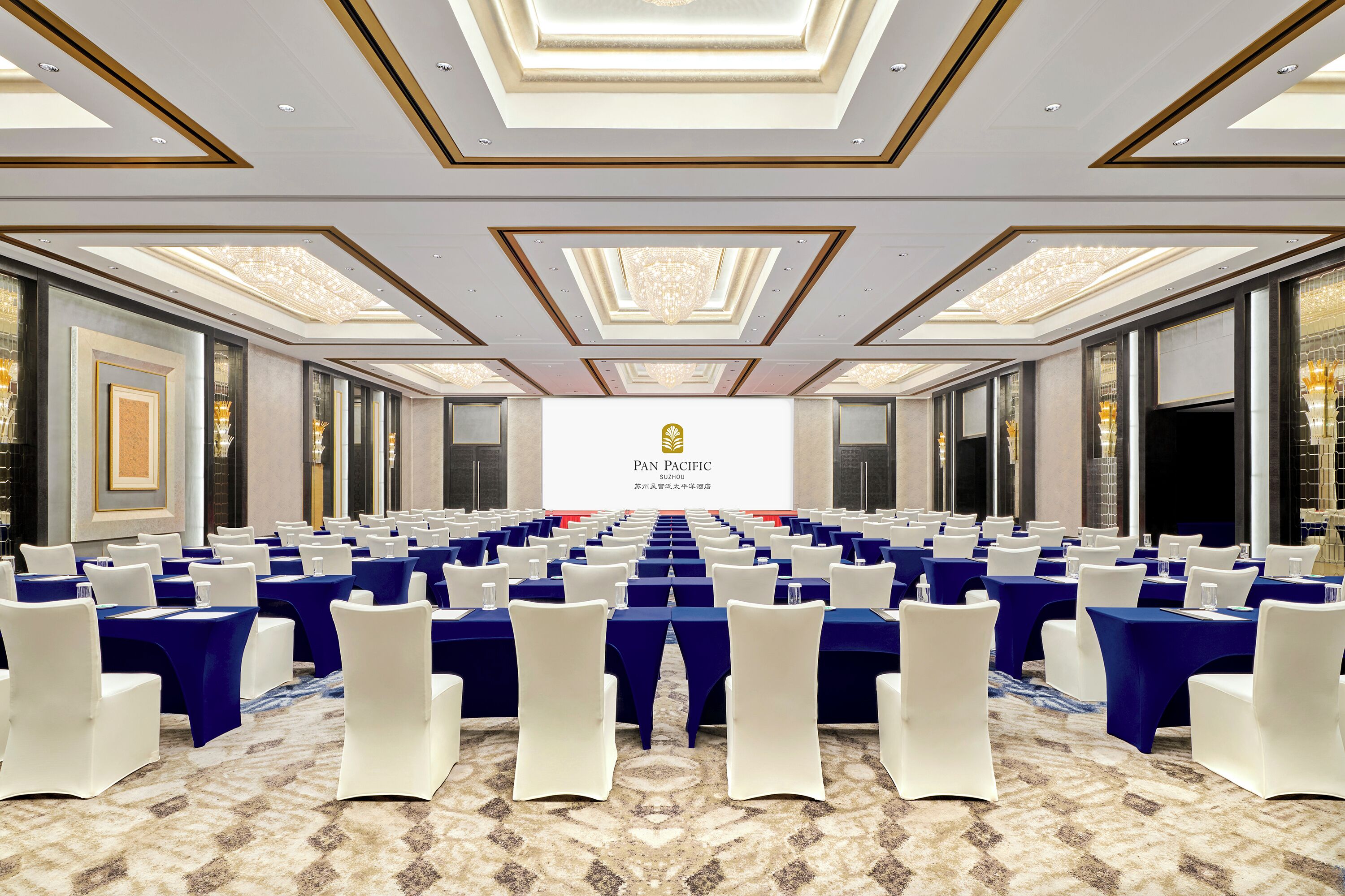 A conference room with rows of tables and chairs, a large screen displaying the logo, and a patterned carpet.