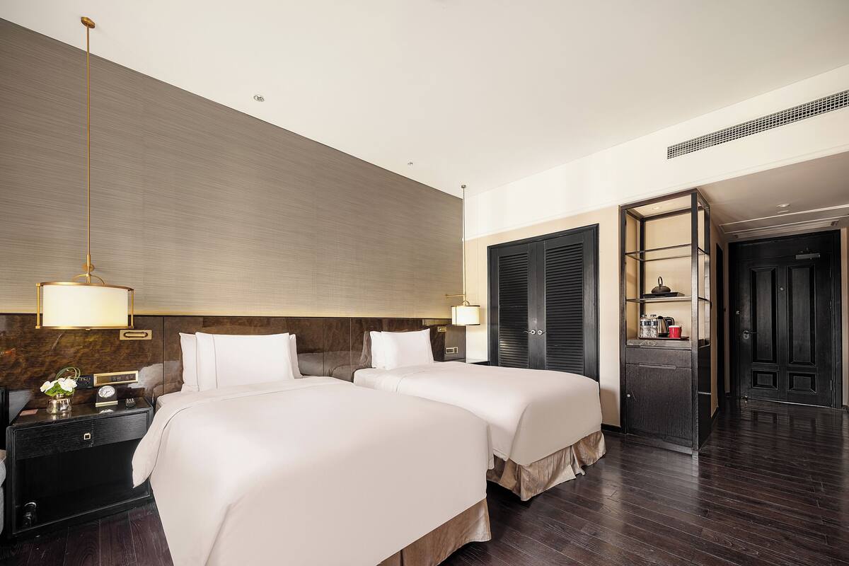 A hotel room with two beds, a nightstand, a lamp, a wardrobe, and a door leading to another room.