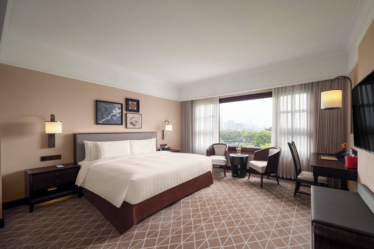 A hotel room with a large bed, a desk with a chair, a small table, and a TV. There are curtains on the window and a lamp on the desk.