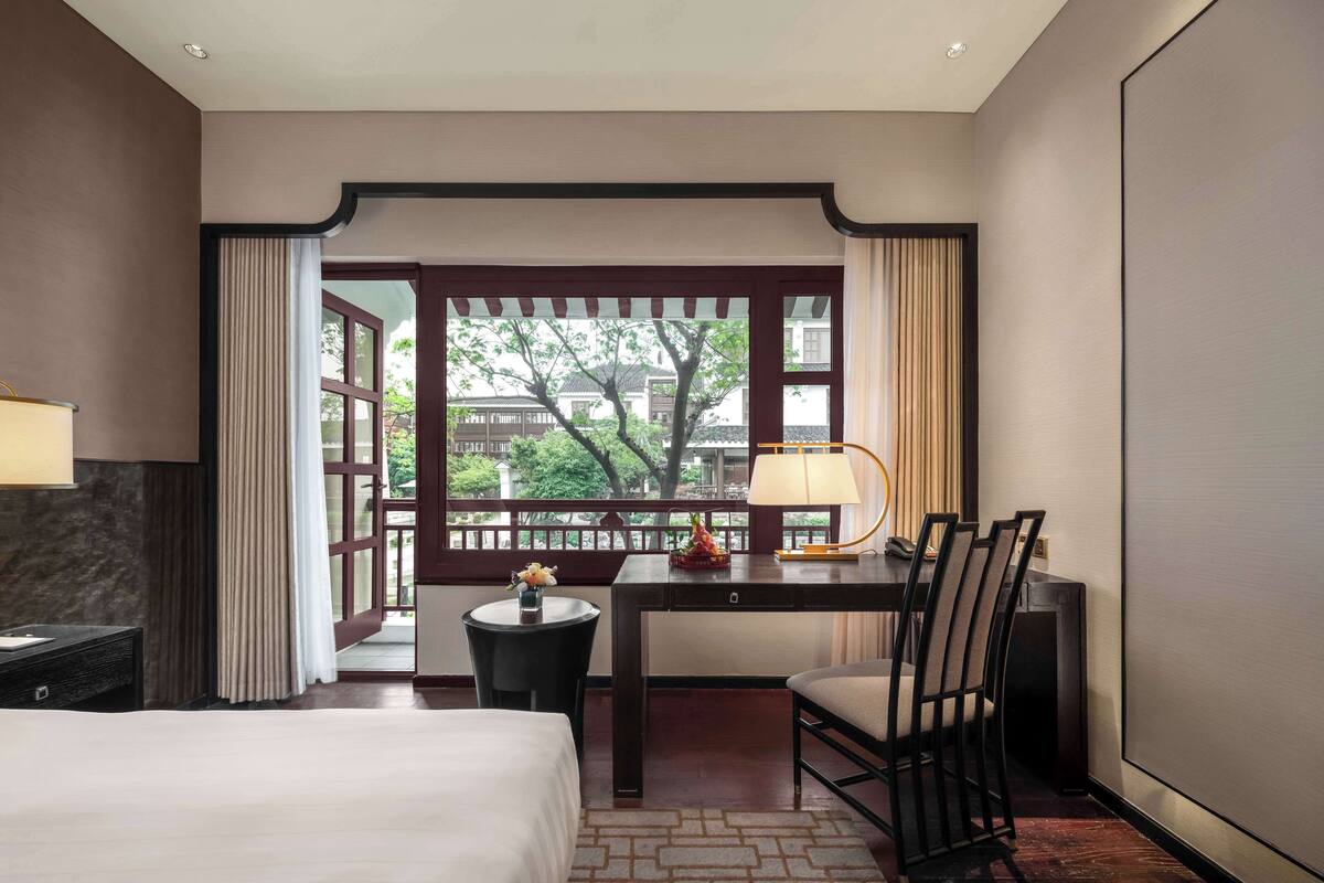 A hotel room with a bed, a desk with a lamp, a chair, and a view through a window with curtains.