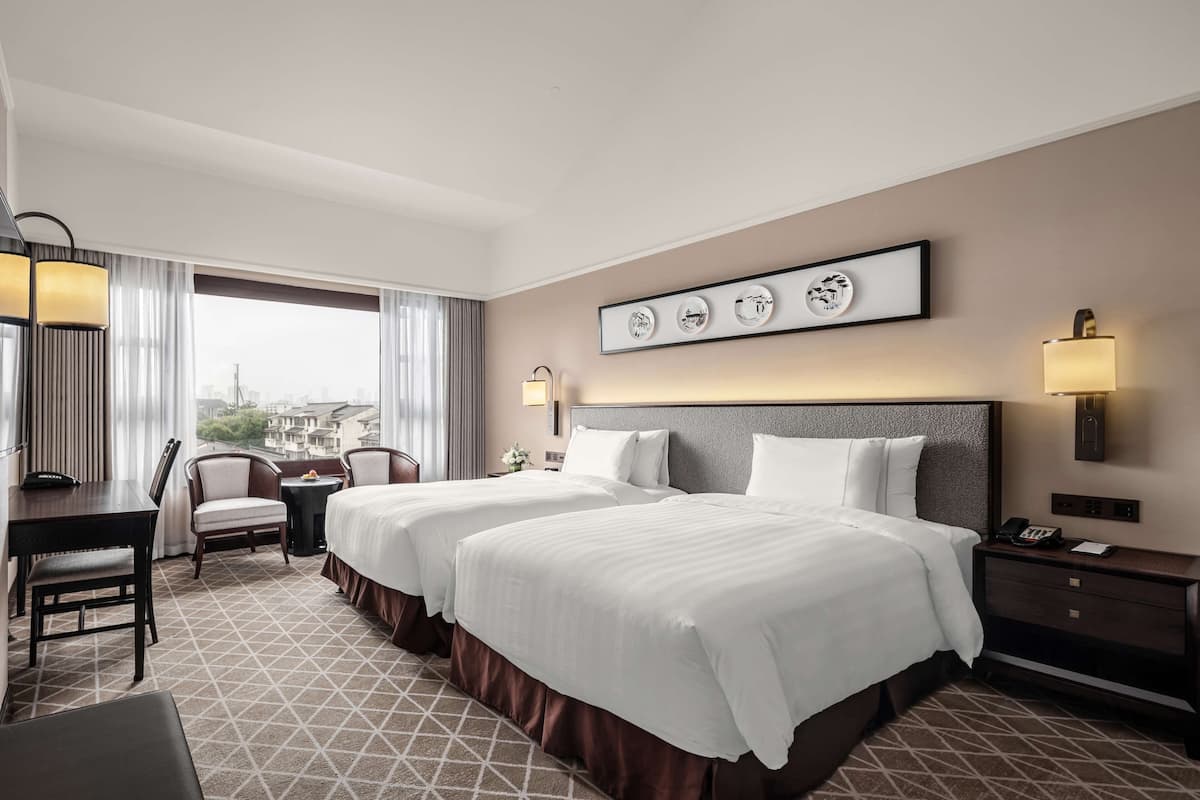 A hotel room with two beds, a desk, a chair, and a view of the city.