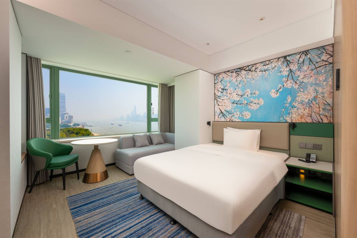 A hotel room with a large bed, a green chair, a gray sofa, a small table, and a view of the city through a large window.