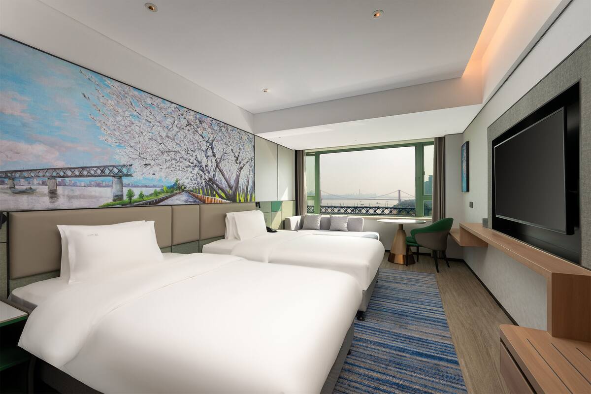 A hotel room with two beds, a TV, a desk, and a painting of a bridge and cherry blossoms.