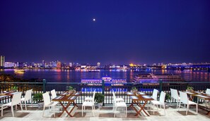 Un restaurant al passeig marítim amb taules i caires, vista a l'horitzó urbà i un riu ple de barques.