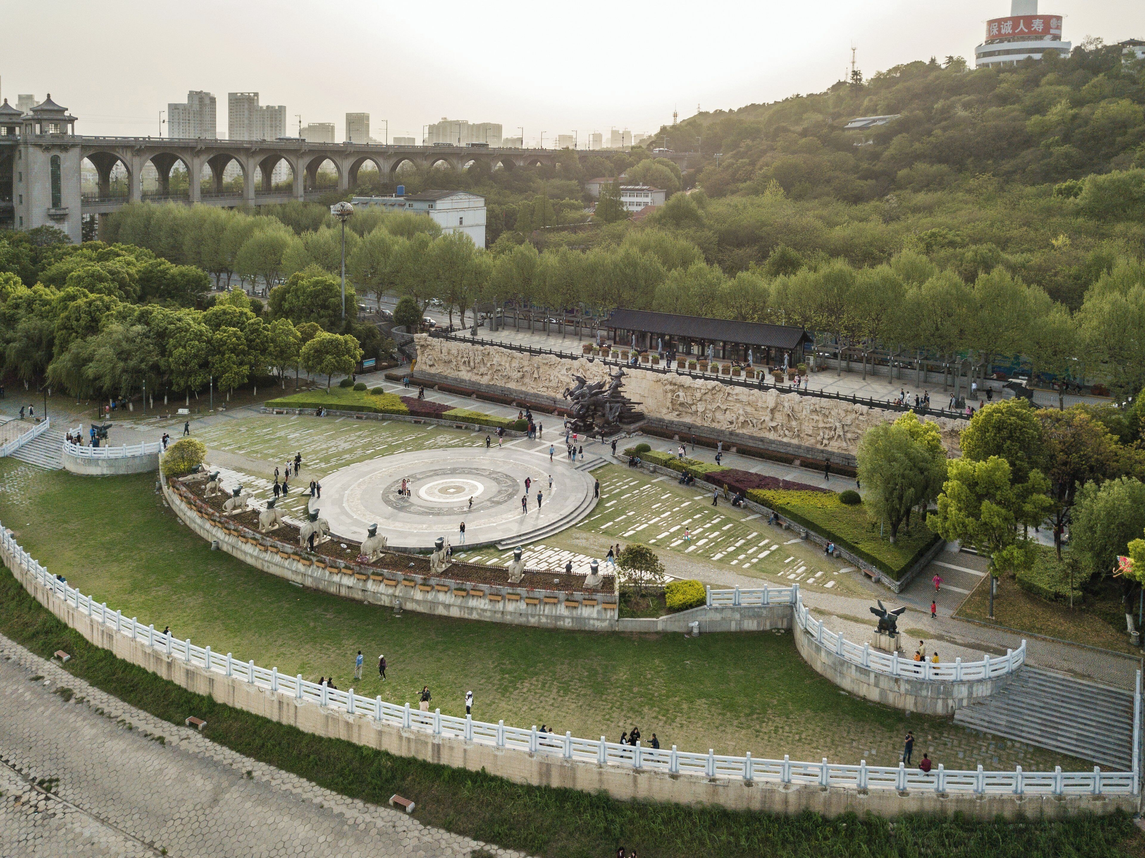 Una amplia plaza abierta con un monumento circular central, rodeado por un muro bajo y una cerca blanca.