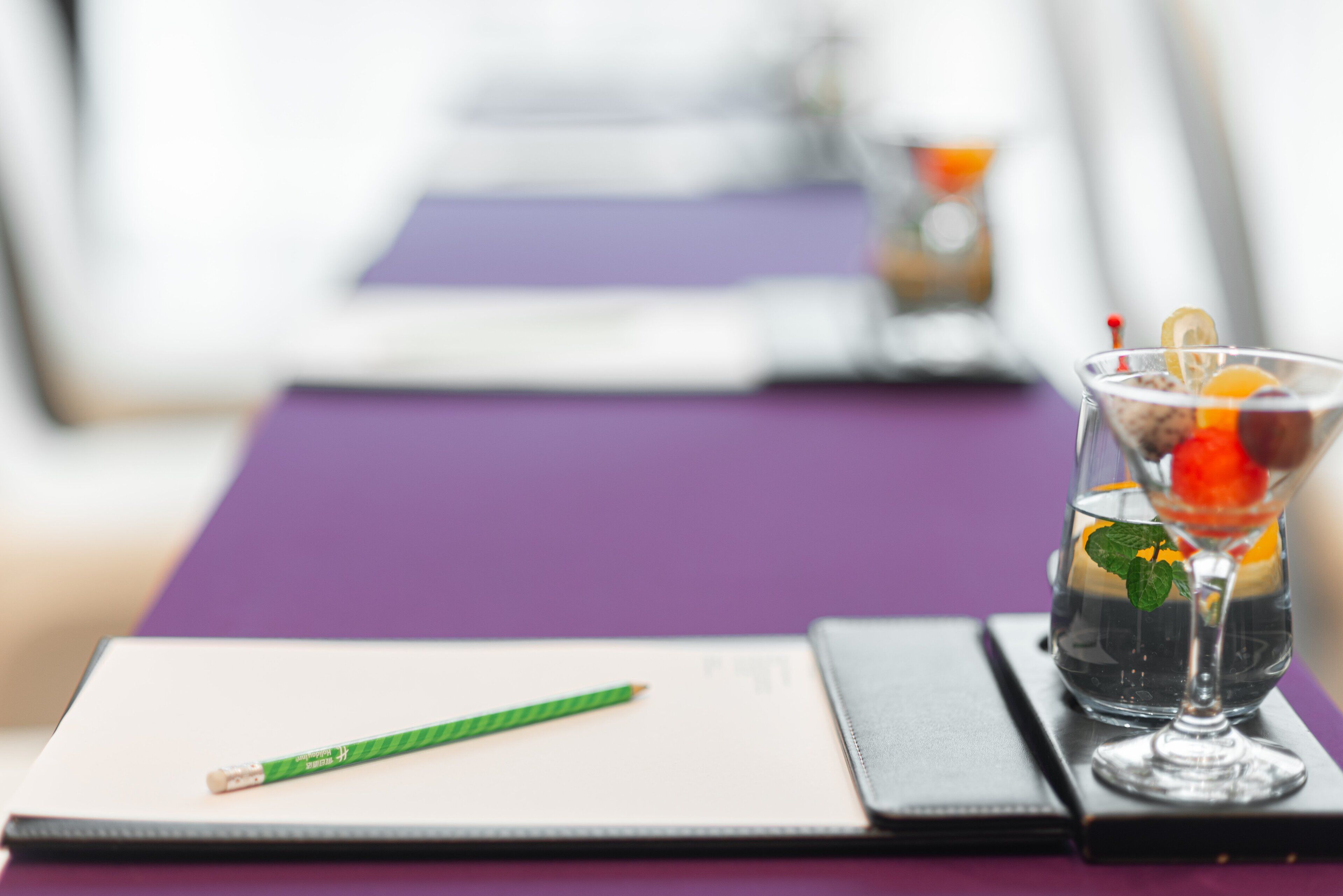 A notebook with a green pencil on a purple table, a glass with a drink and a straw, and a blurred background.