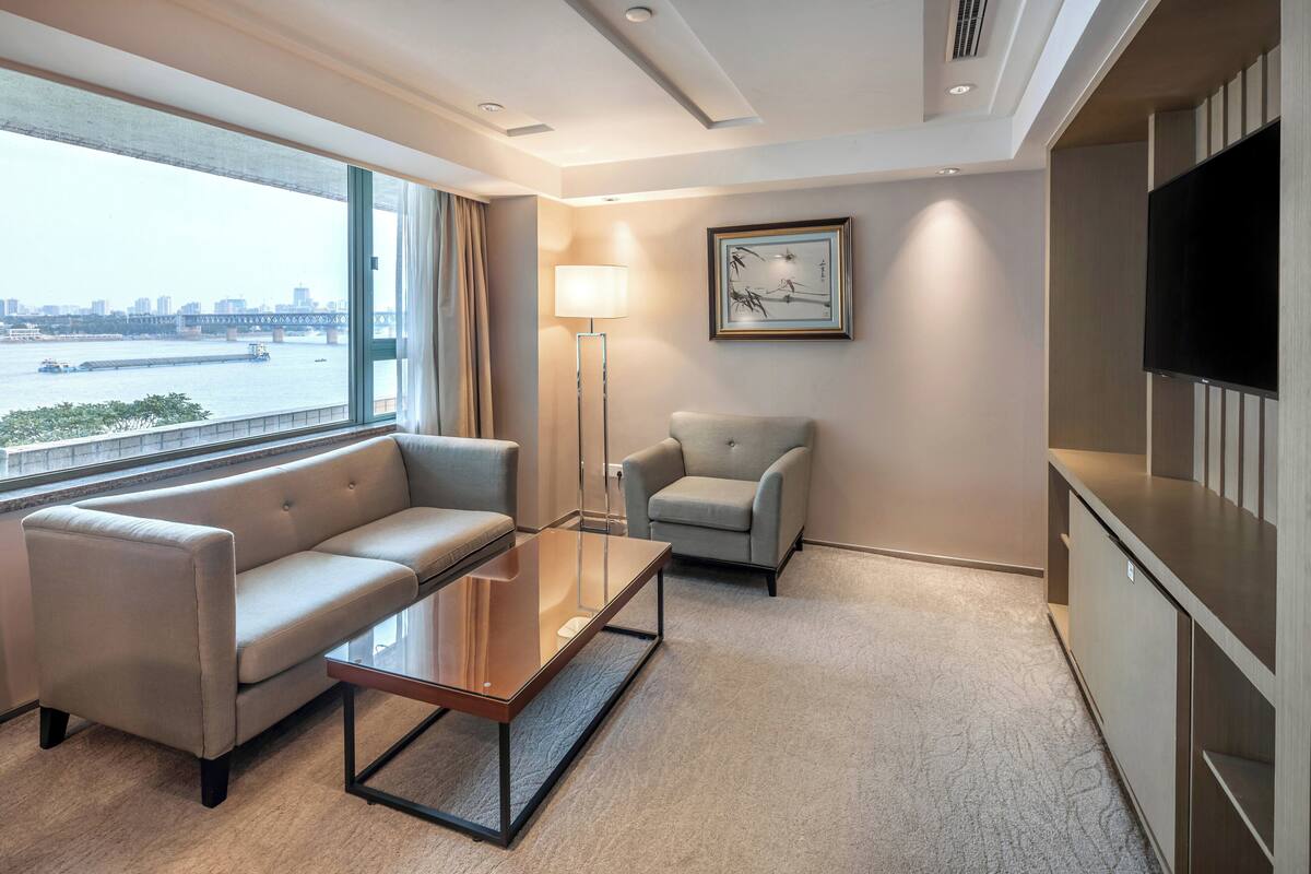 A modern hotel room with a sofa, armchair, flat-screen TV, and a large window offering a view of a river and city skyline.