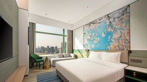 A modern hotel room with a large bed, a desk, a chair, and a view of the city skyline.