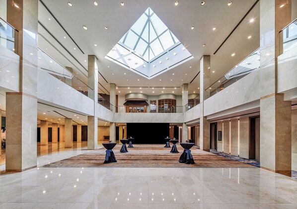 A spacious atrium with a high ceiling, a central open area with tables, and a glass ceiling allowing natural light.