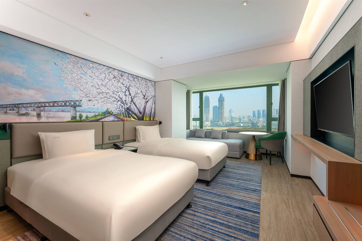 A hotel room with two beds, a large TV, a painting of a bridge, and a view of the city skyline.