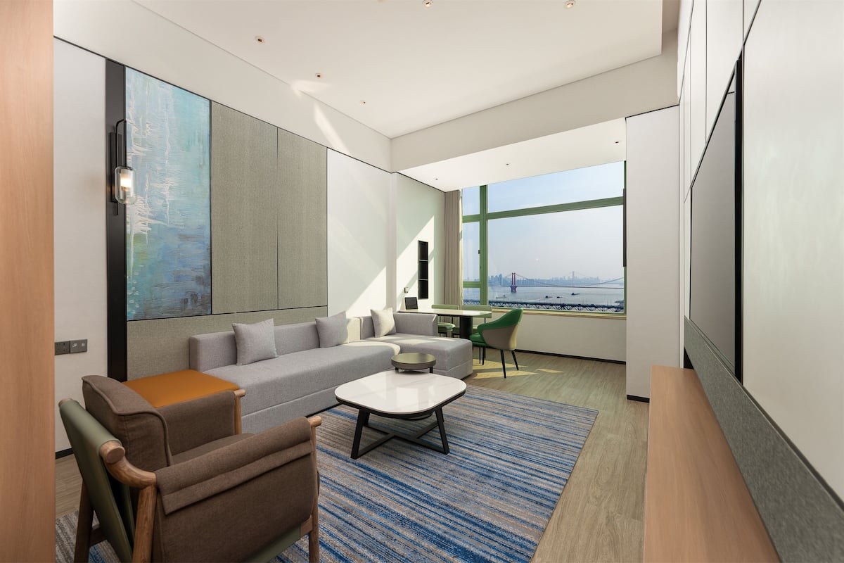 A modern living room with a sofa, armchair, coffee table, and a large window offering a city view.