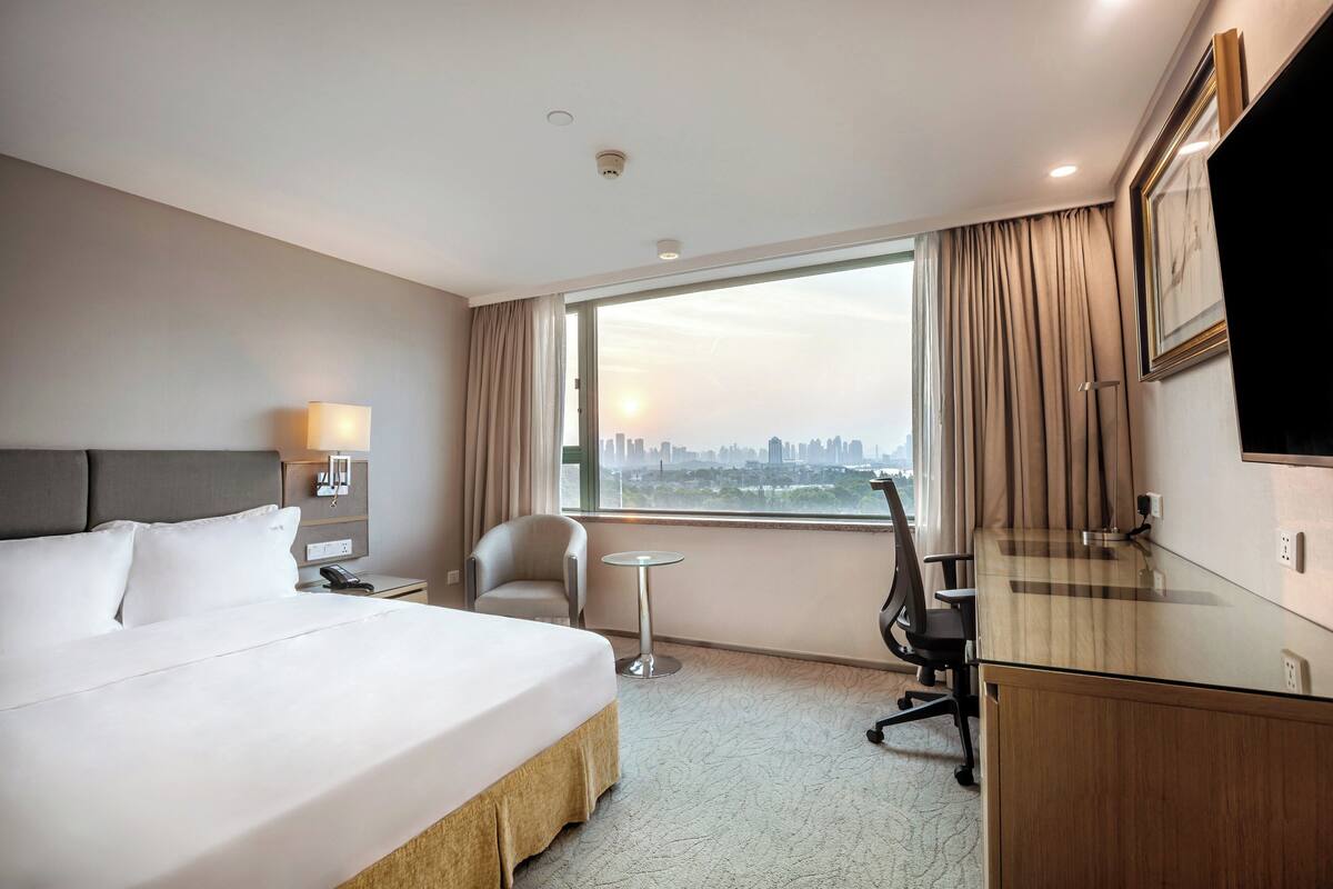 A hotel room with a large bed, a desk, a chair, a small table, and a city view through the window.