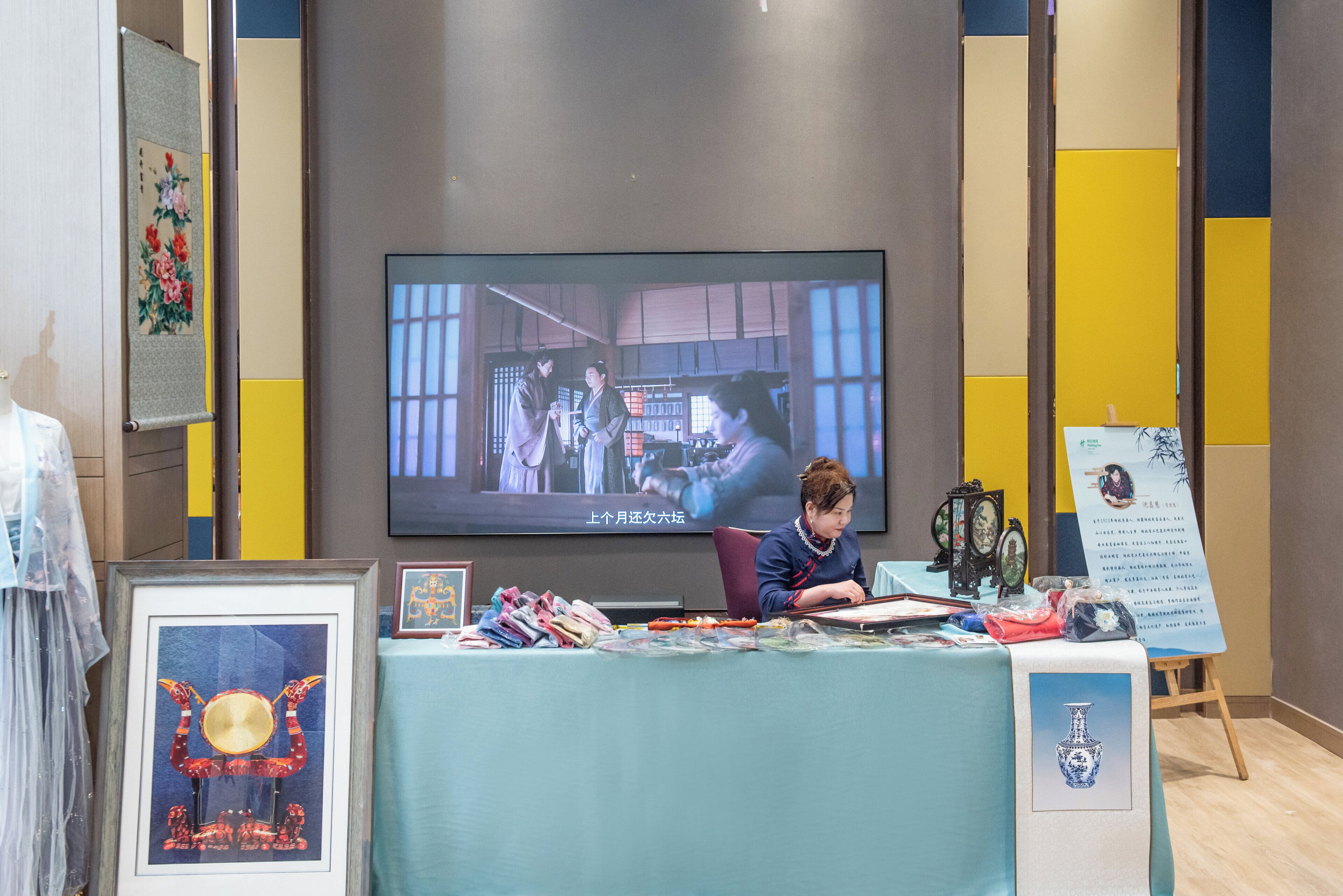 Quarto com televisão mostrando uma cena, mesa com diversos objetos e obras de arte emolduradas nas paredes.