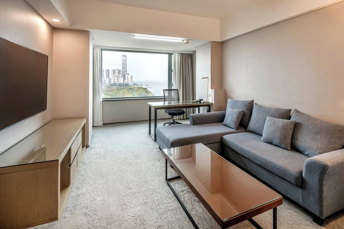 A modern hotel room with a sofa, a TV, a desk, and a view of a cityscape.