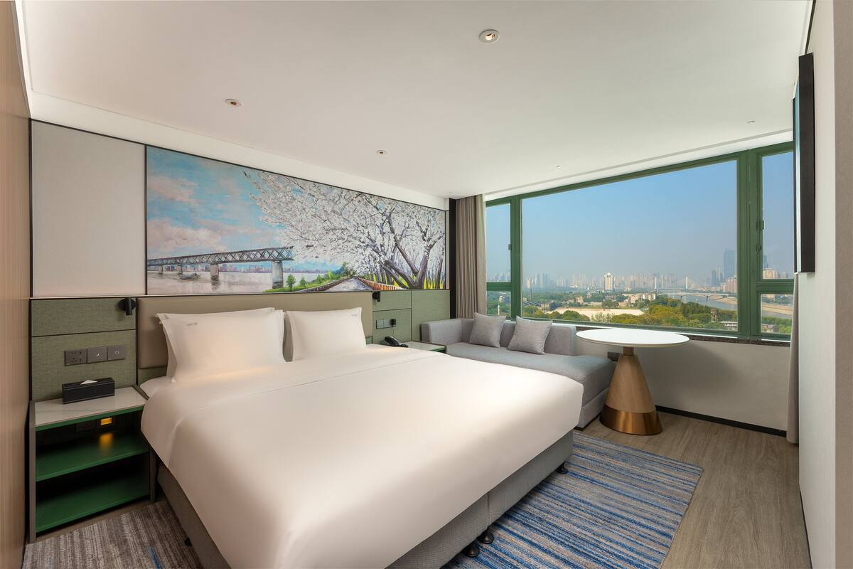 A modern hotel room with a large bed, a sofa, a small table, and a city view through a large window.