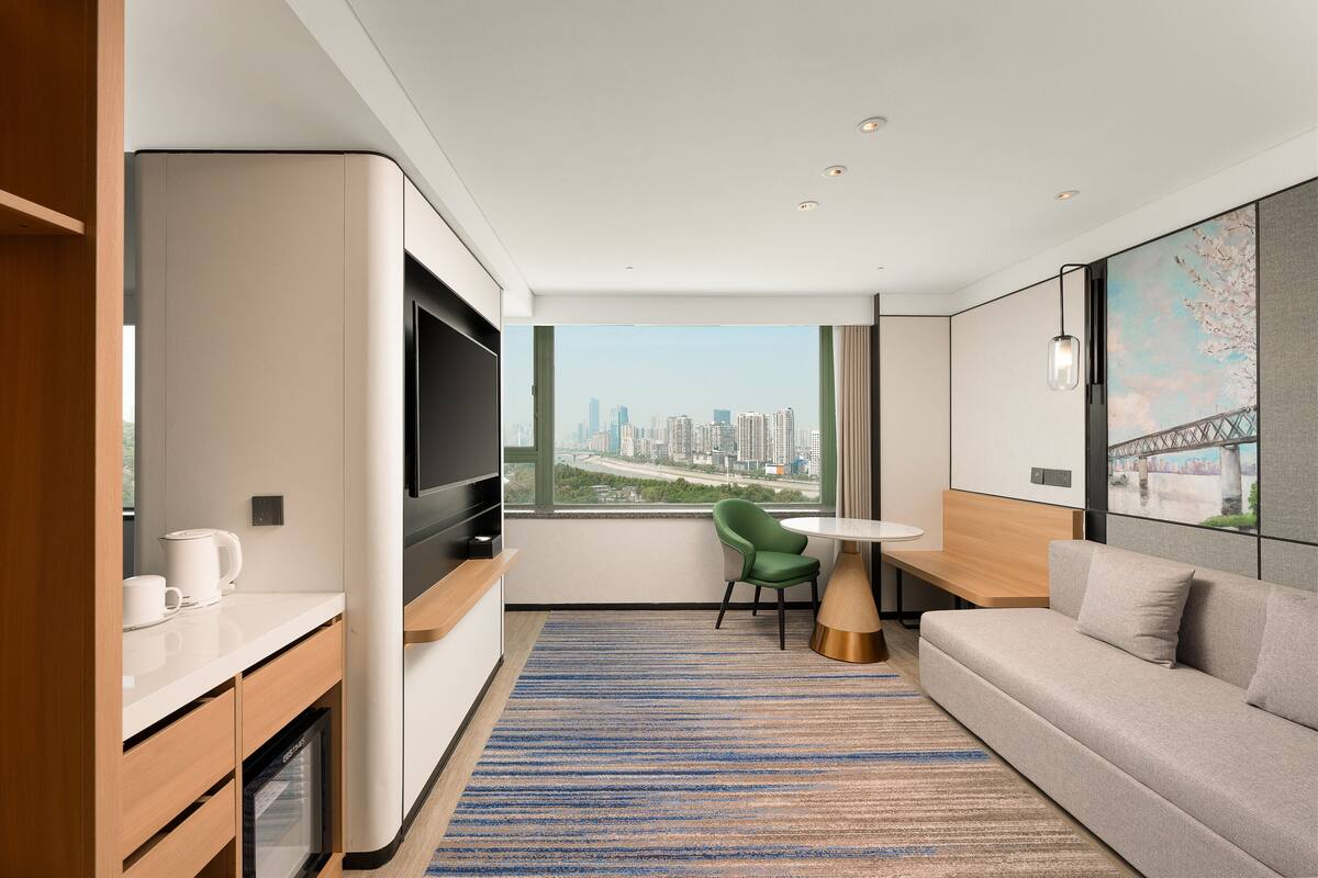 A modern hotel room with a city view, a sofa, a green chair, a small table, and a TV.