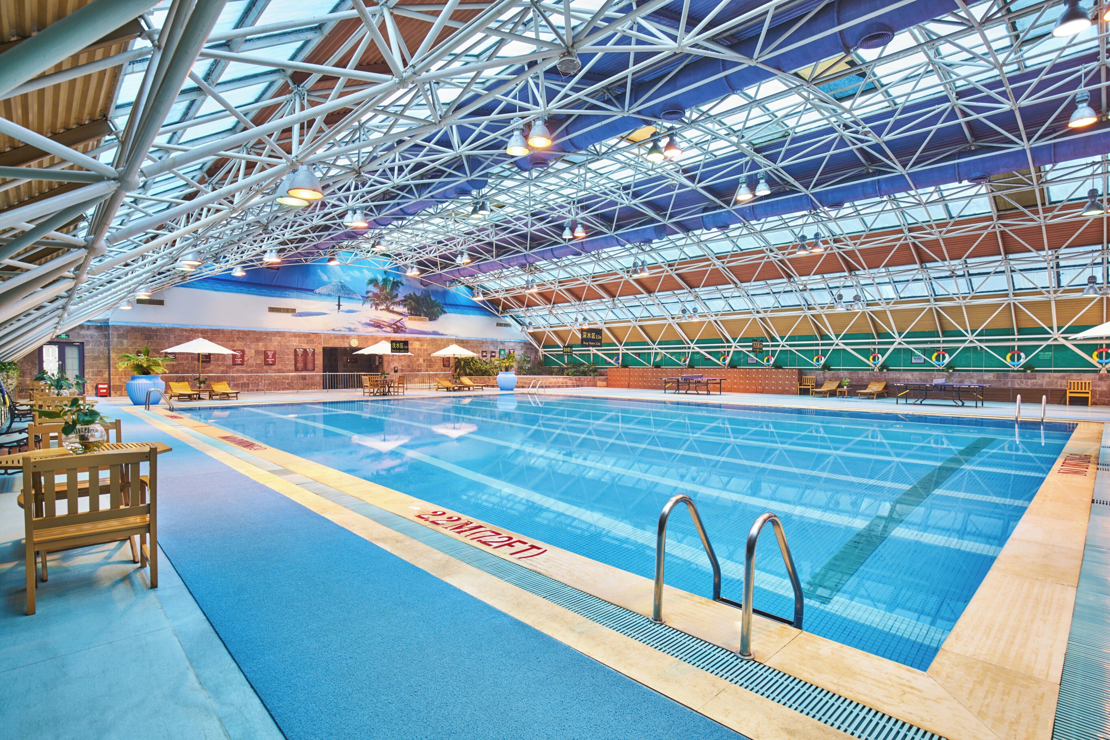 An indoor swimming pool with a high ceiling and metal framework, surrounded by lounge chairs and tables.