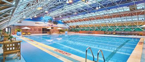 An indoor swimming pool with a high ceiling and metal framework, surrounded by lounge chairs and tables.