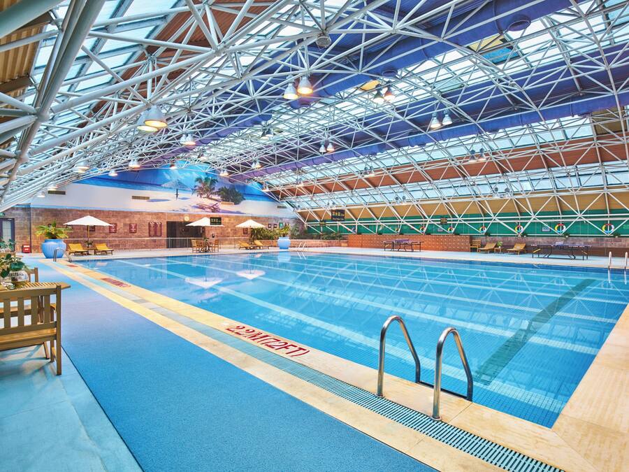 An indoor swimming pool with a high ceiling and metal framework, surrounded by lounge chairs and tables.