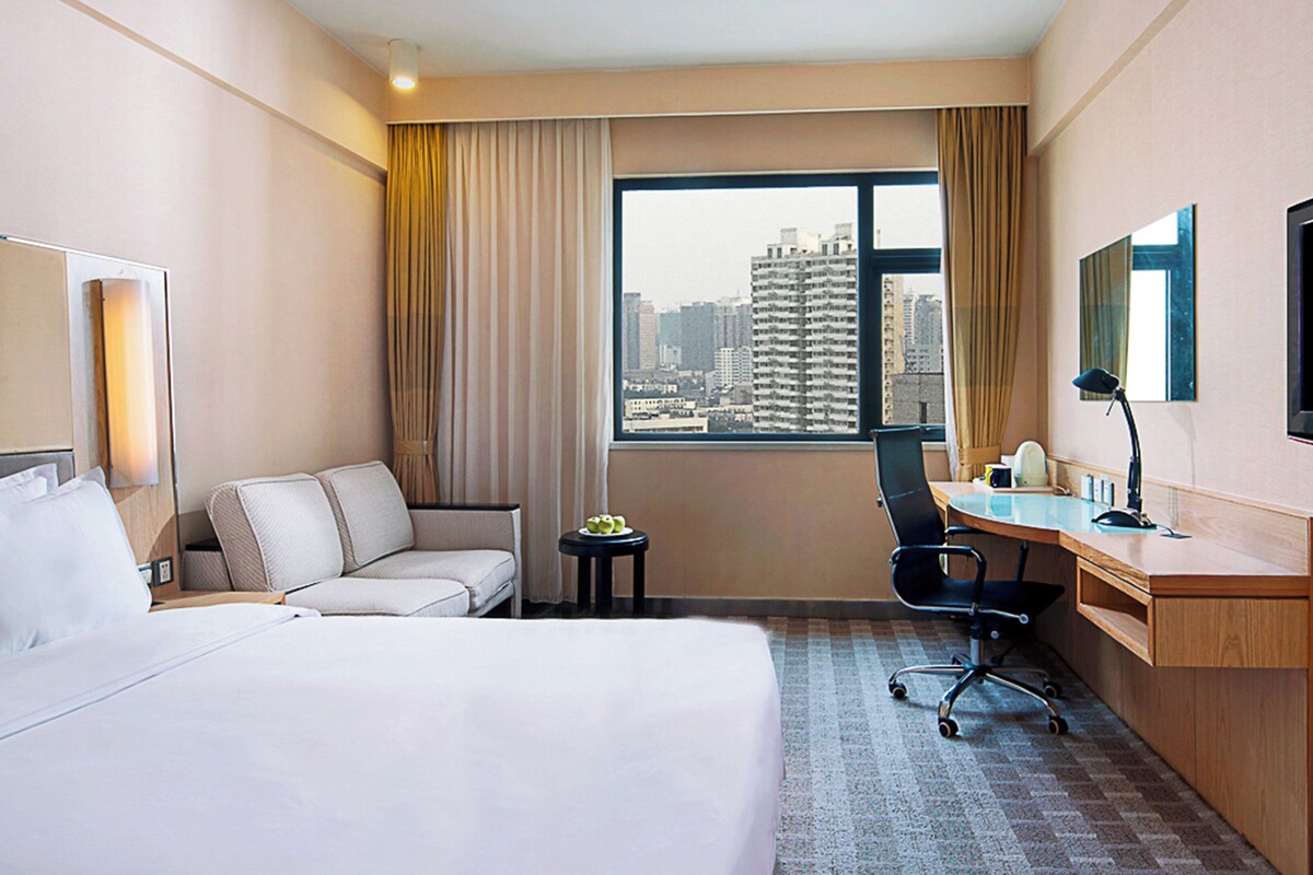 A hotel room with a bed, a desk, a chair, a television, and a window with a city view.