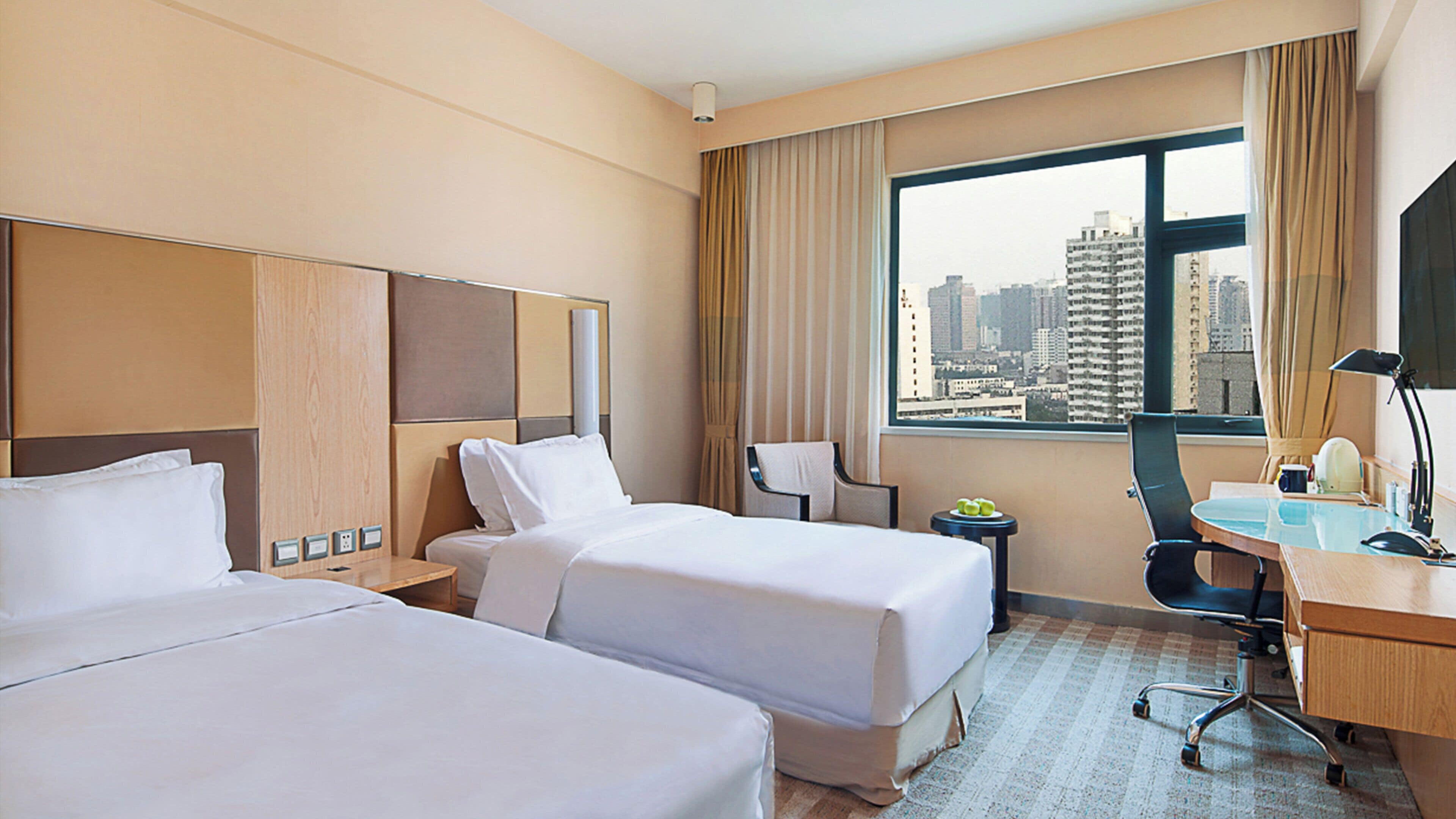 A hotel room with two beds, a desk, a chair, a lamp, and a window with a city view.