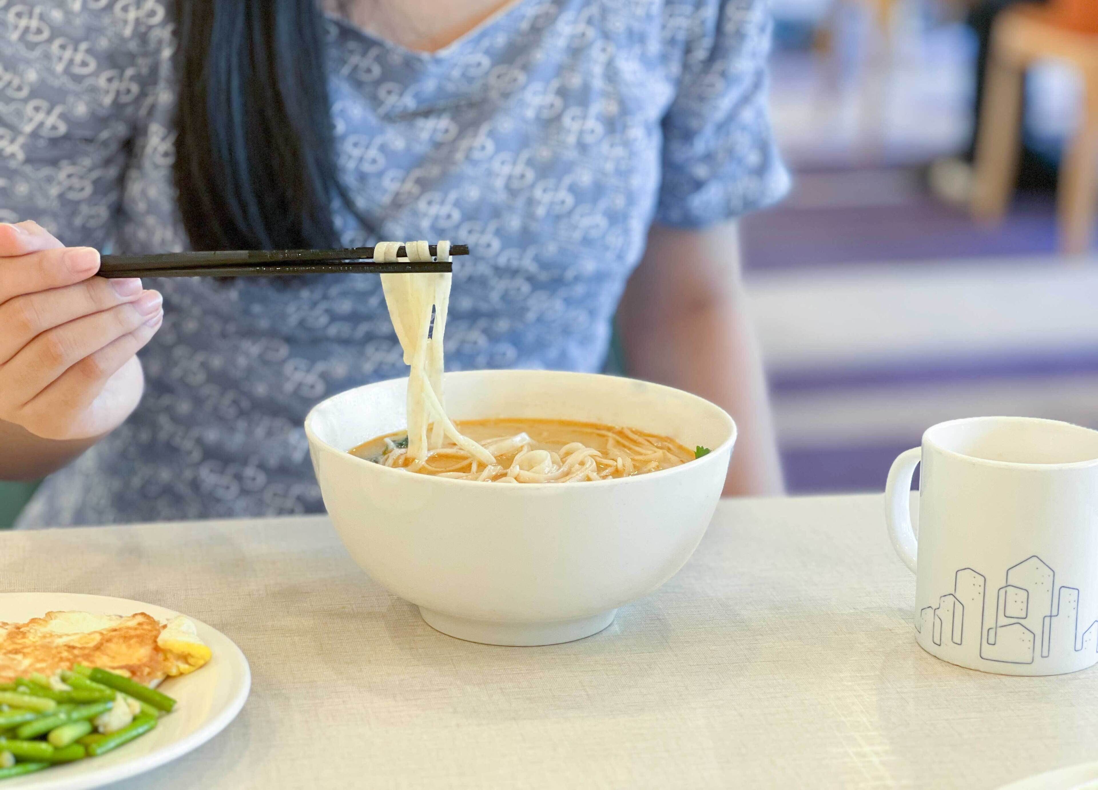 A person eating noodles with chopsticks, a white bowl, and a white mug on a table.