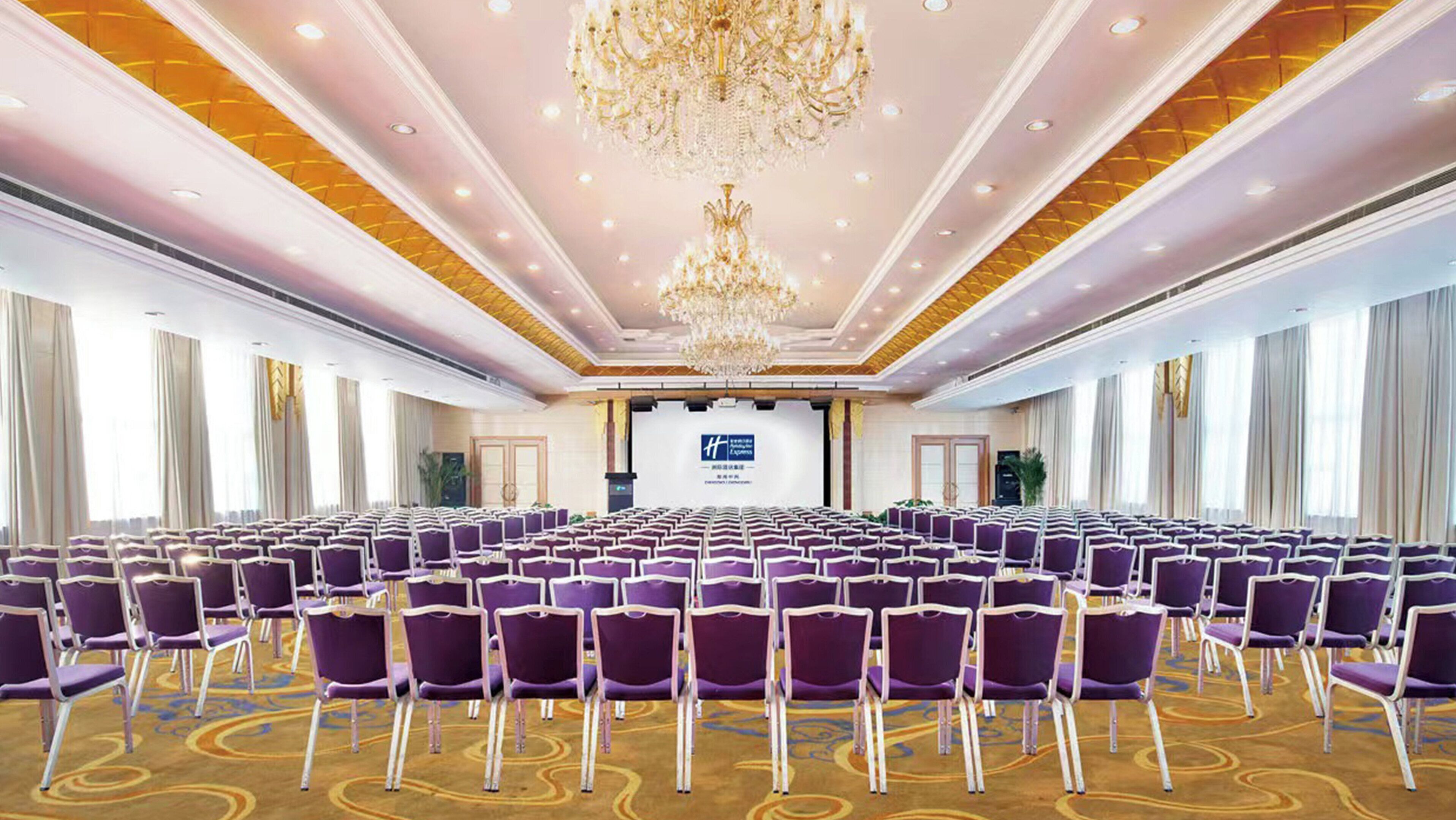 Ein Konferenzraum mit Reihen violettfarbener Stühle, einer großen Kristallleuchter und einer Bühne mit einem Bildschirm im Vordergrund.