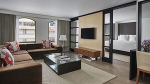 A modern hotel room with a sofa, a coffee table, a TV, a bed, and a large window.