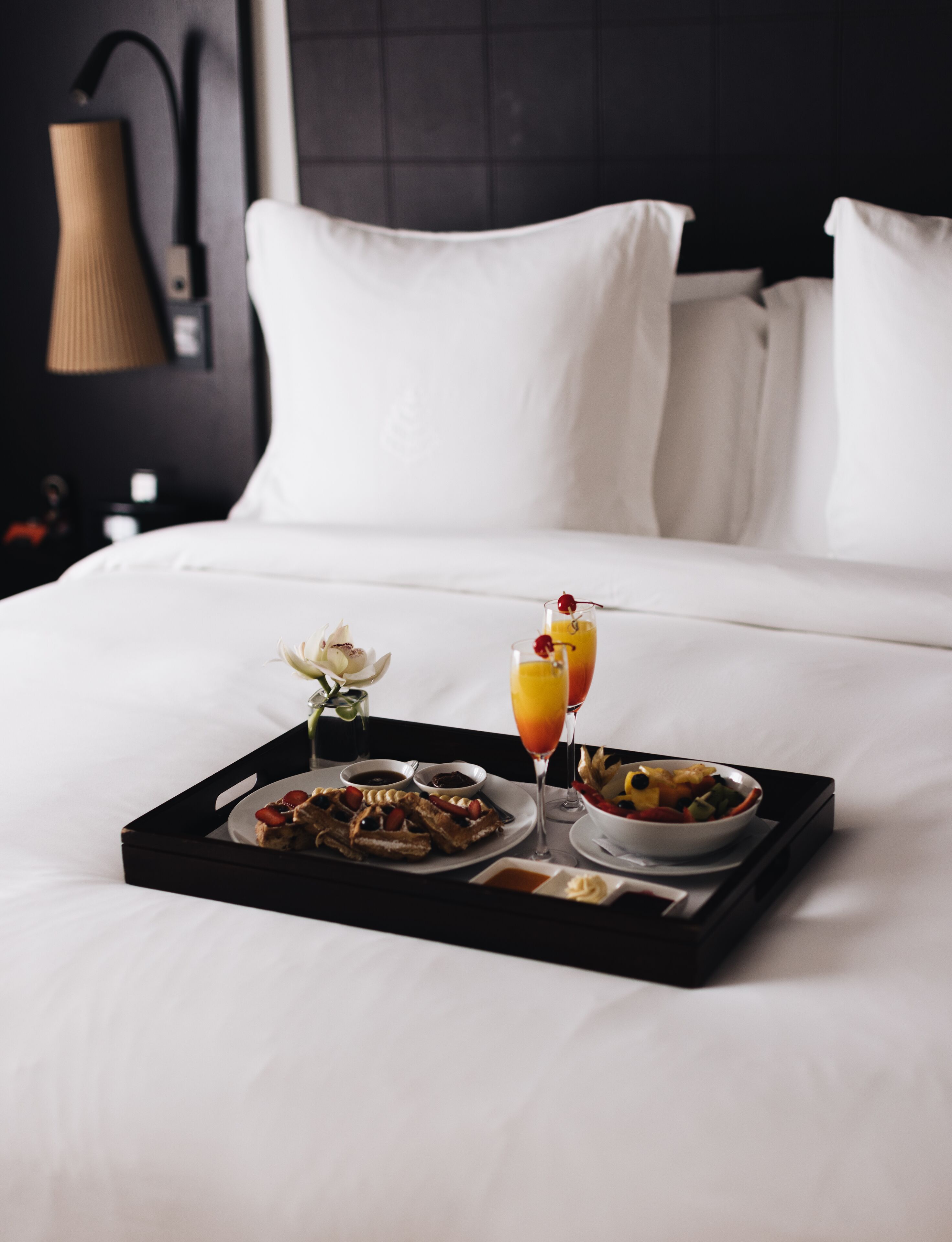 A neatly made breakfast tray on a bed with waffles, fruit, and two drinks, in a room with white bedding and a dark headboard.