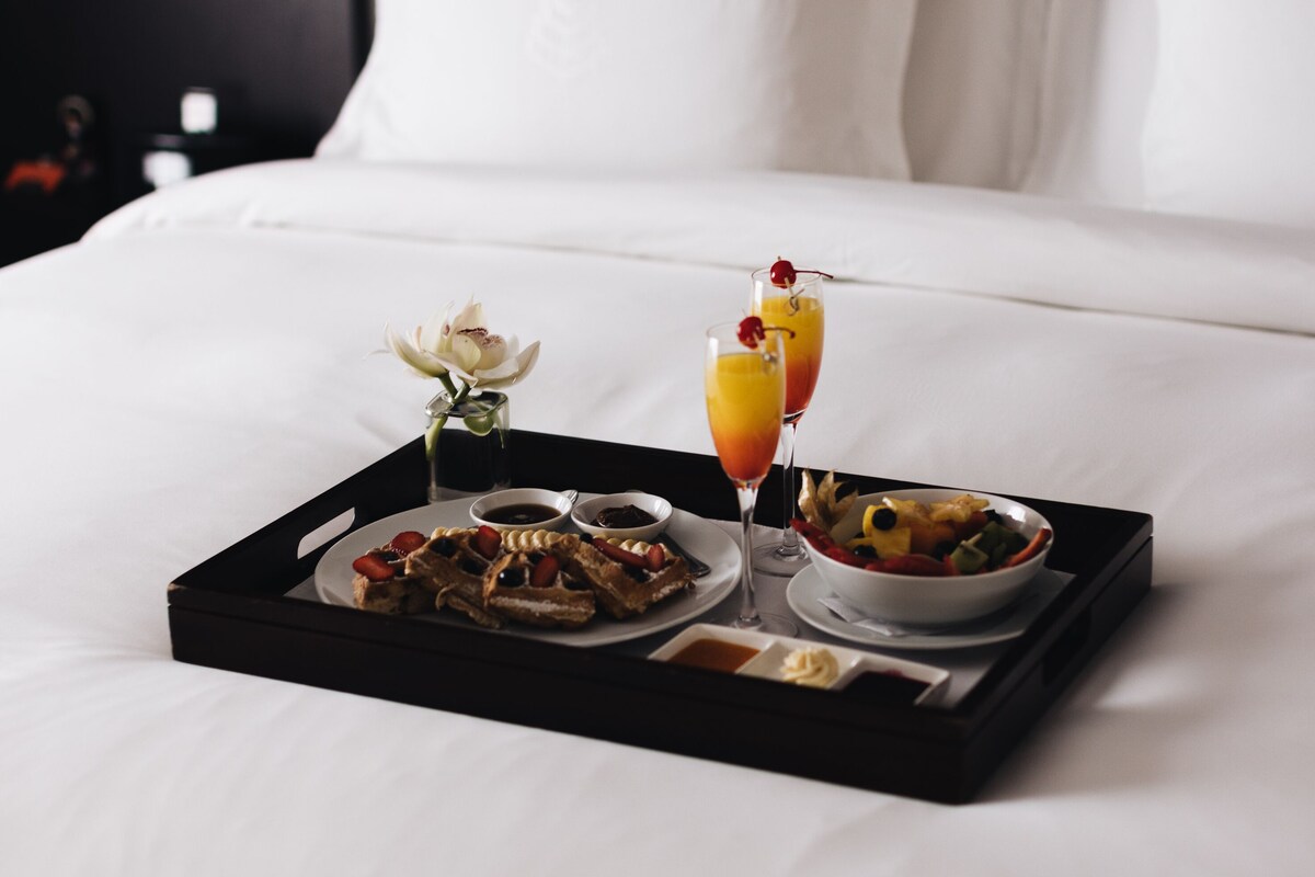 A neatly made breakfast tray on a bed with waffles, fruit, and two drinks, in a room with white bedding and a dark headboard.
