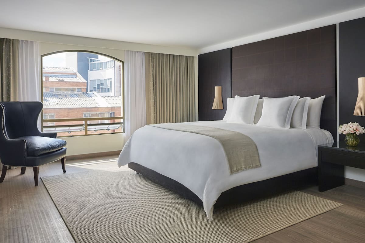 A modern hotel room with a large bed, a chair, a nightstand, and a view of buildings through the window.