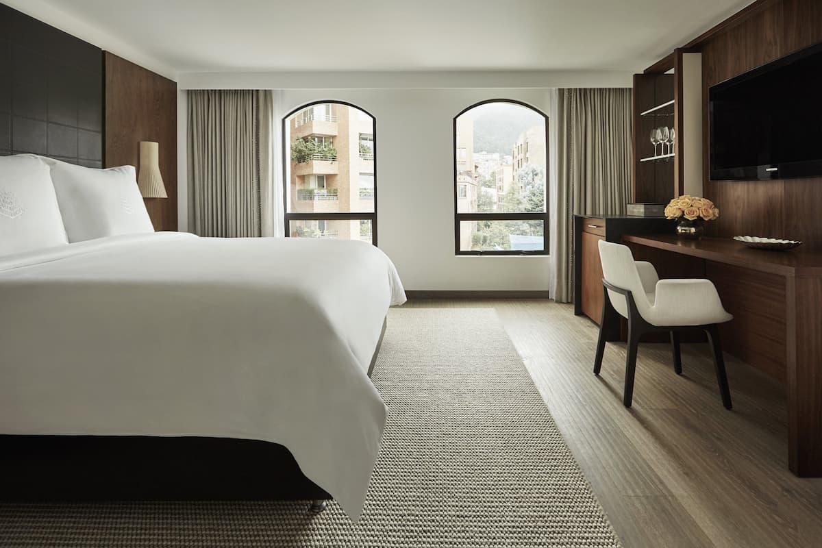 A hotel room with a large bed, a desk with a chair, a TV, and a view of buildings through arched windows.