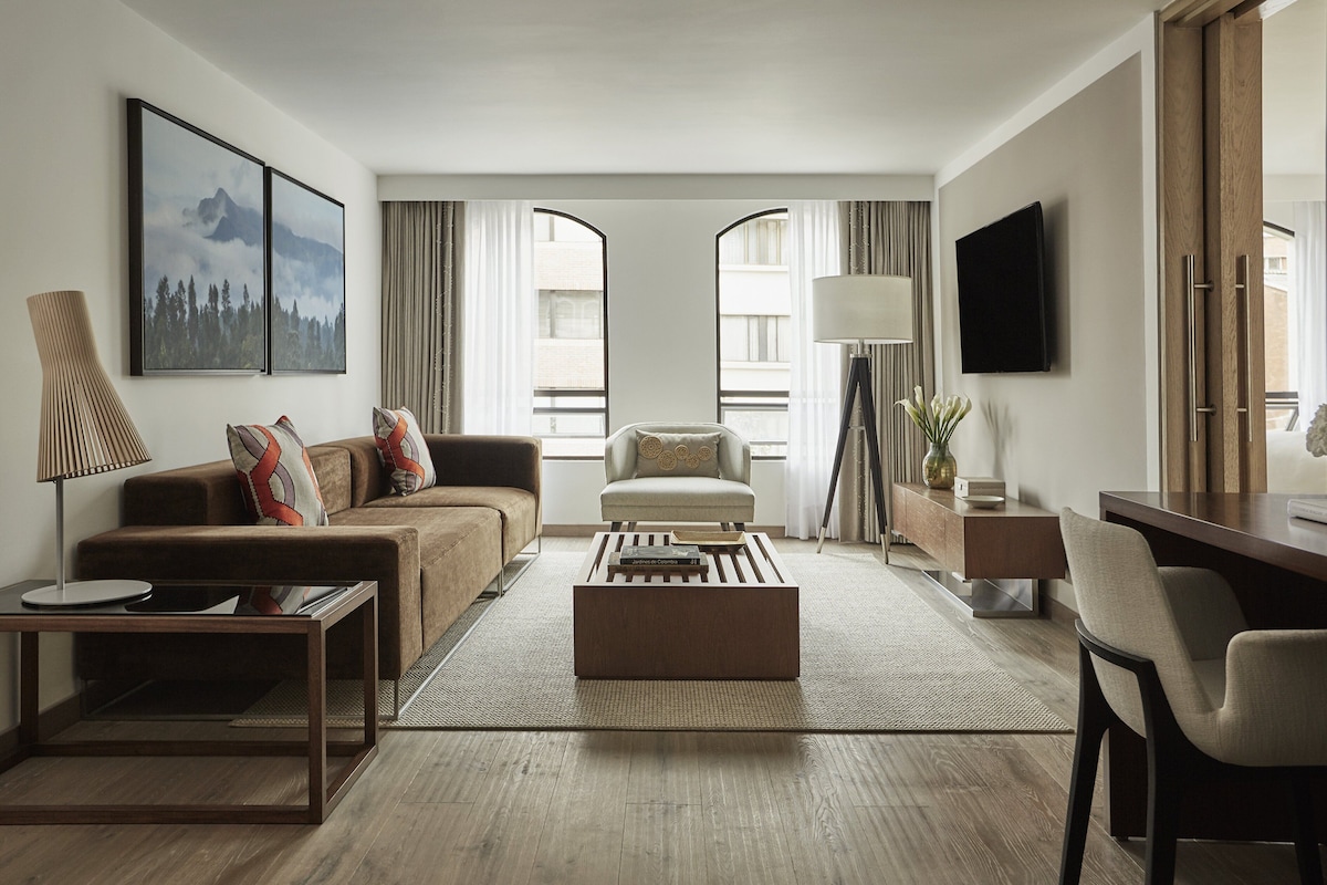 Habitación de hotel moderna con sofá, sillón, mesa de centro y escritorio con silla. Cuenta con dos ventanas amplias con cortinas, un televisor de pantalla plana montado en la pared y una lámpara sobre una mesita de noche.