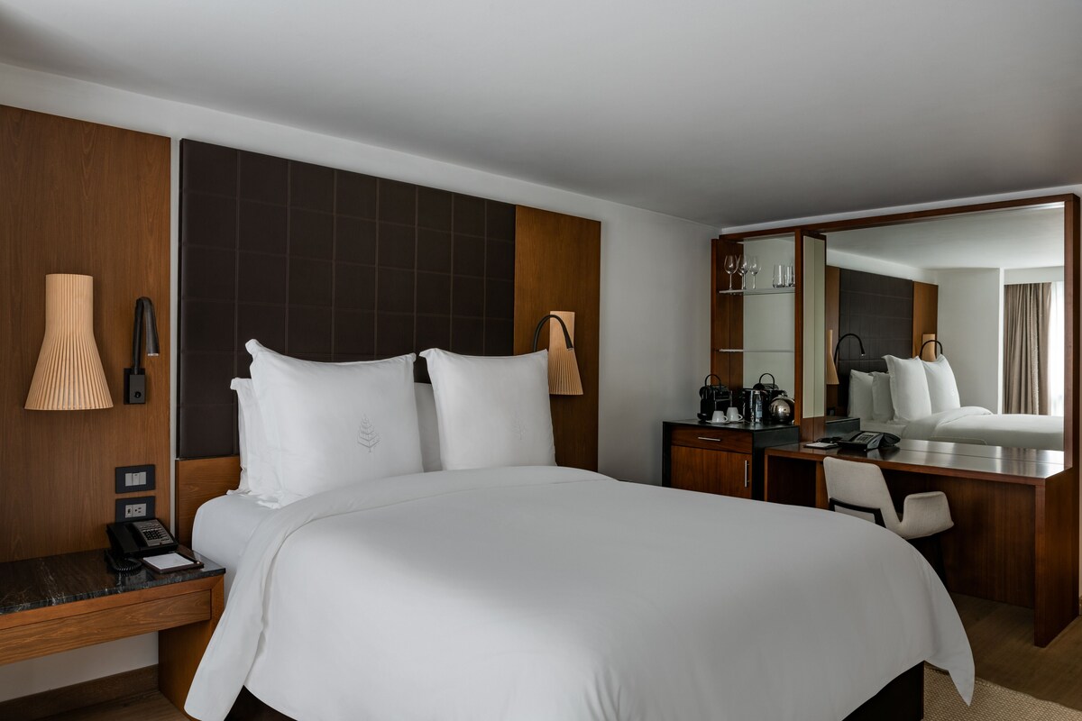 A hotel room with a large bed, a desk, a chair, a telephone, a lamp, and a mirror.