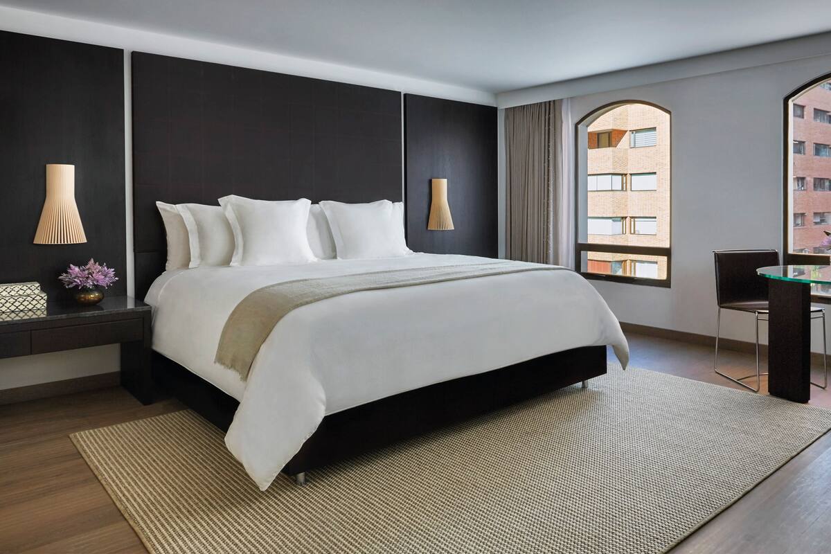 A modern hotel room with a large bed, a desk with chairs, and a view of a building through a window.
