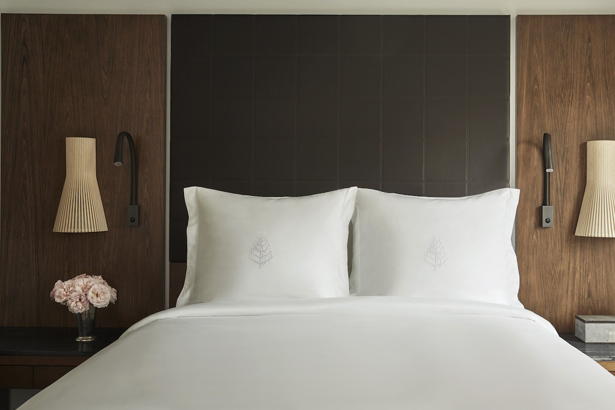 Habitación de hotel con una cama grande, dos almohadas, un florero con flores y dos lámparas de pared.
