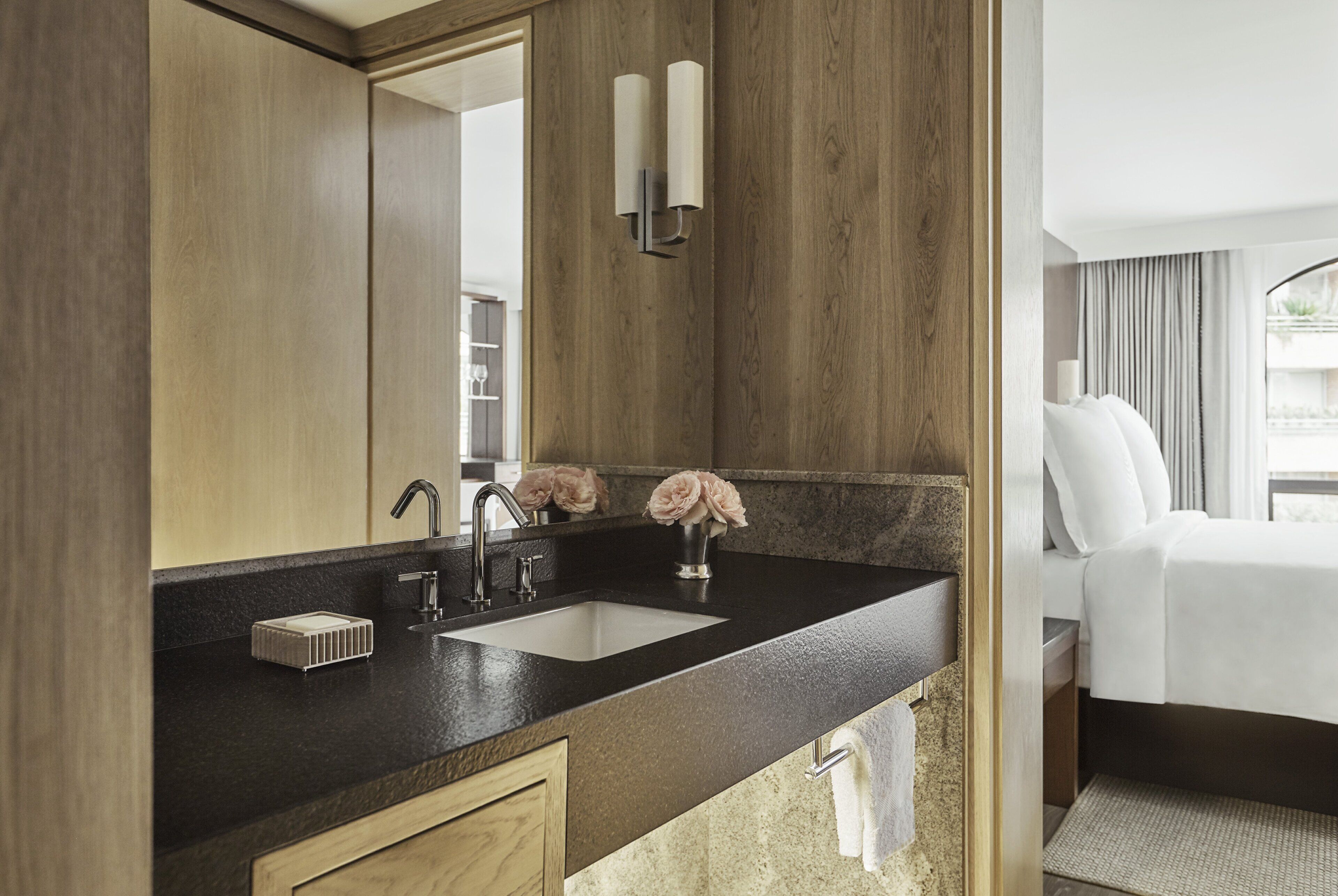Een moderne badkamer met een grote spiegel, twee wasbakken, een wastafelmeubel, en een bed dat door een open deur zichtbaar is.