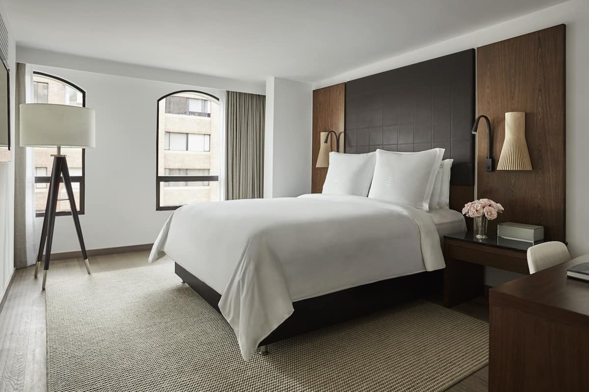 A modern hotel room with a large bed, a desk, a lamp, and a view of the city through a window.